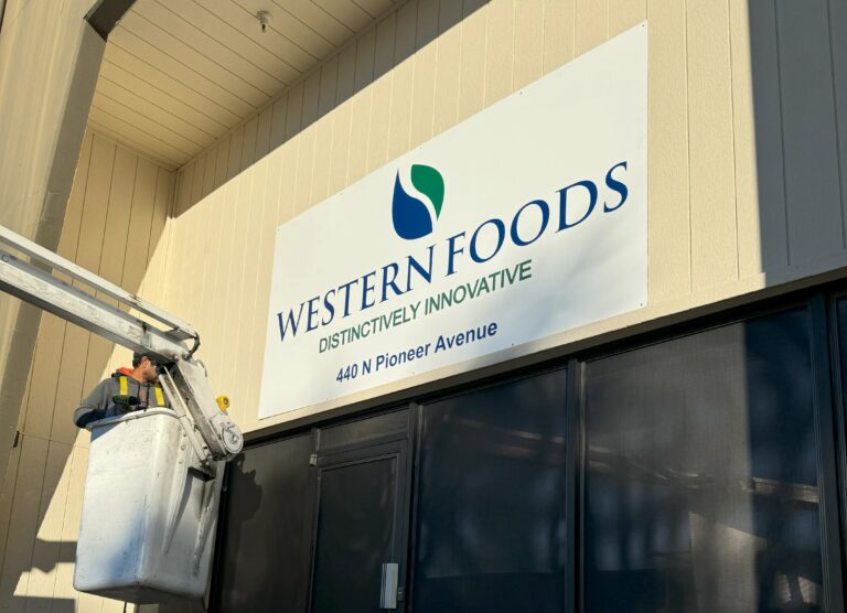 Worker on a lift installing or inspecting a large exterior sign for Western Foods on a beige building.